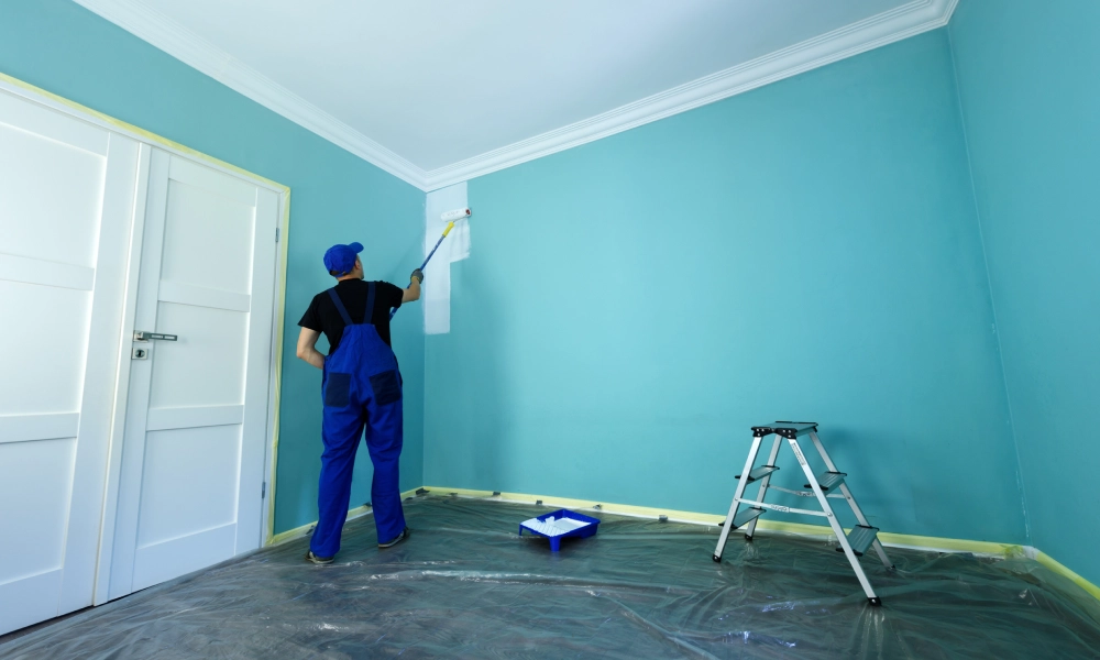 Painter applying white paint on a light blue wall with a roller.