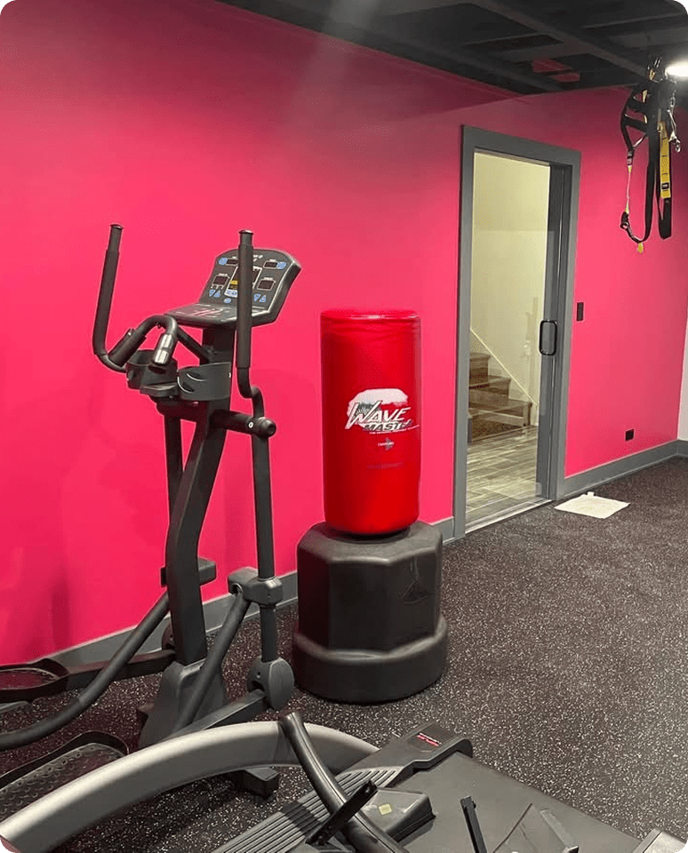 A red punching bag and elliptical machine in a gym with bright pink walls.